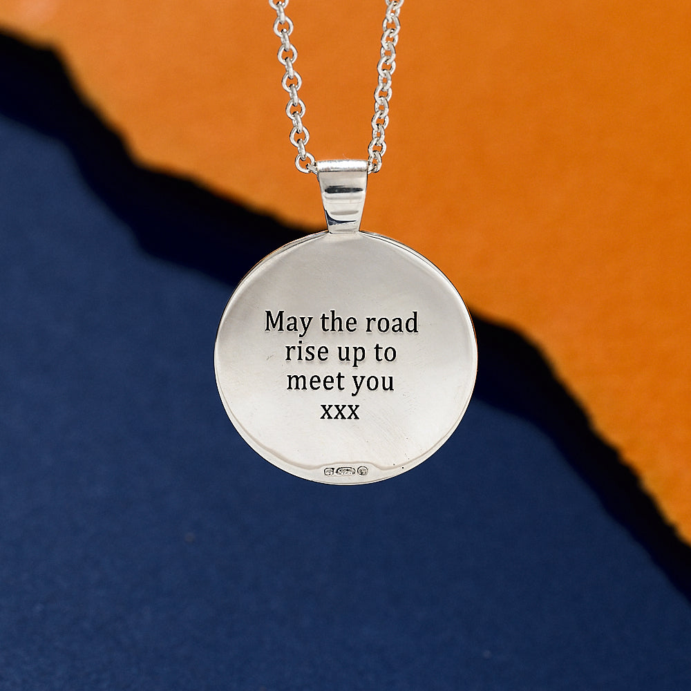 Silver necklace with a round pendant featuring engraved text may the road rise up to meet you