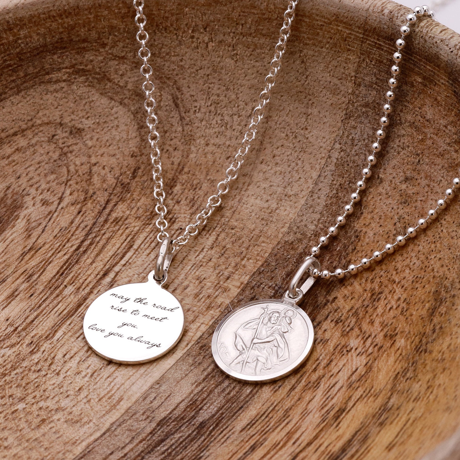 Two silver Saint Christopher necklaces with engraved pendants on a wooden surface