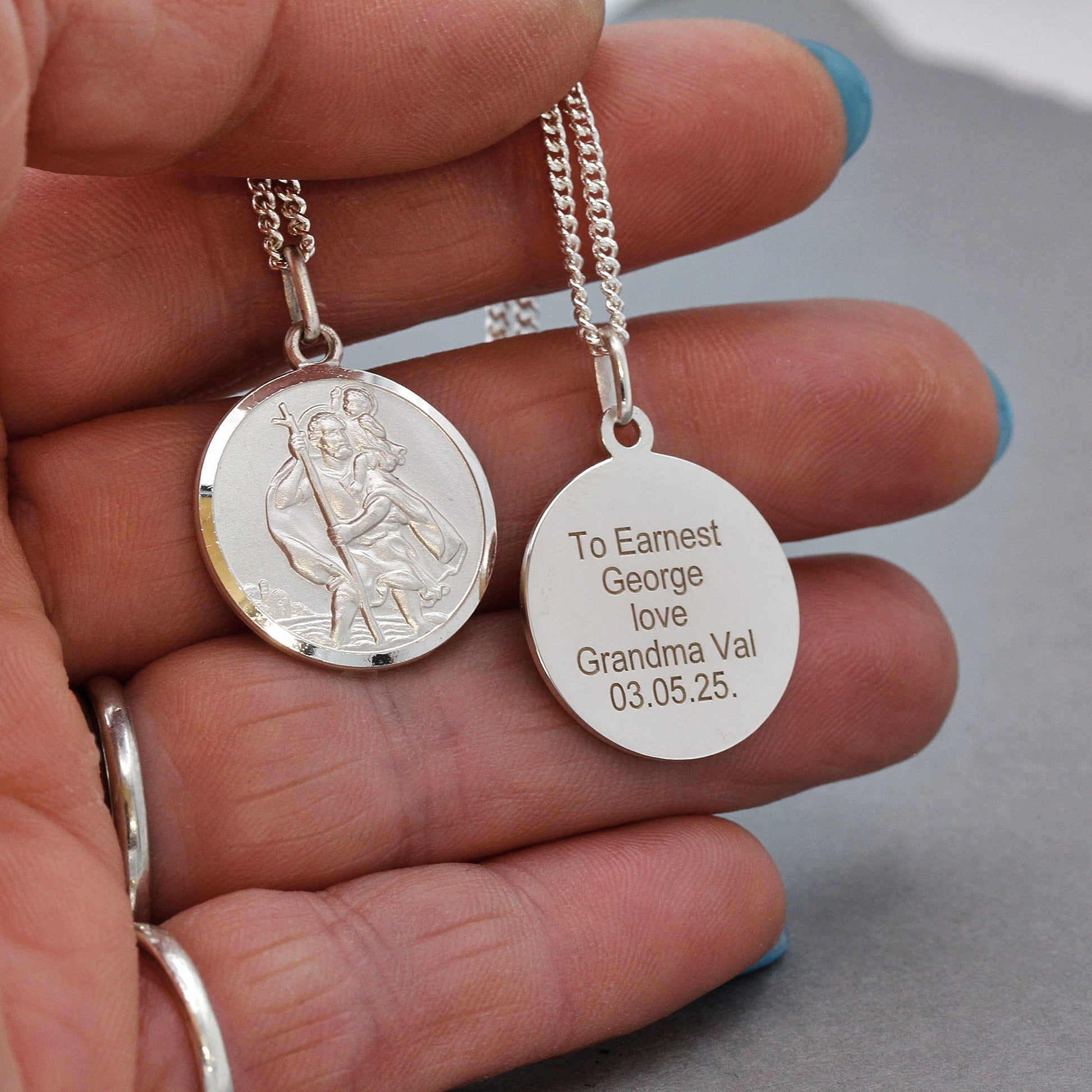 Two silver pendants held in a hand, one with an engraved message and the other with a religious design.