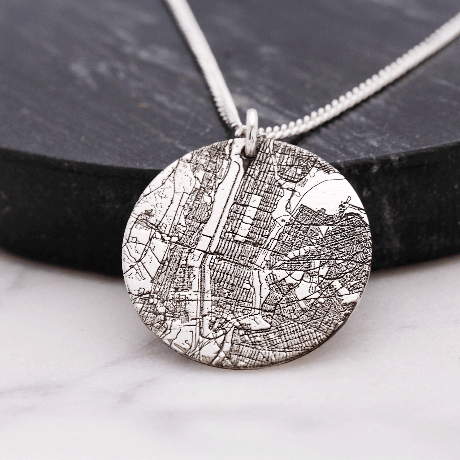 Silver necklace with a round pendant featuring a map design on a dark surface.