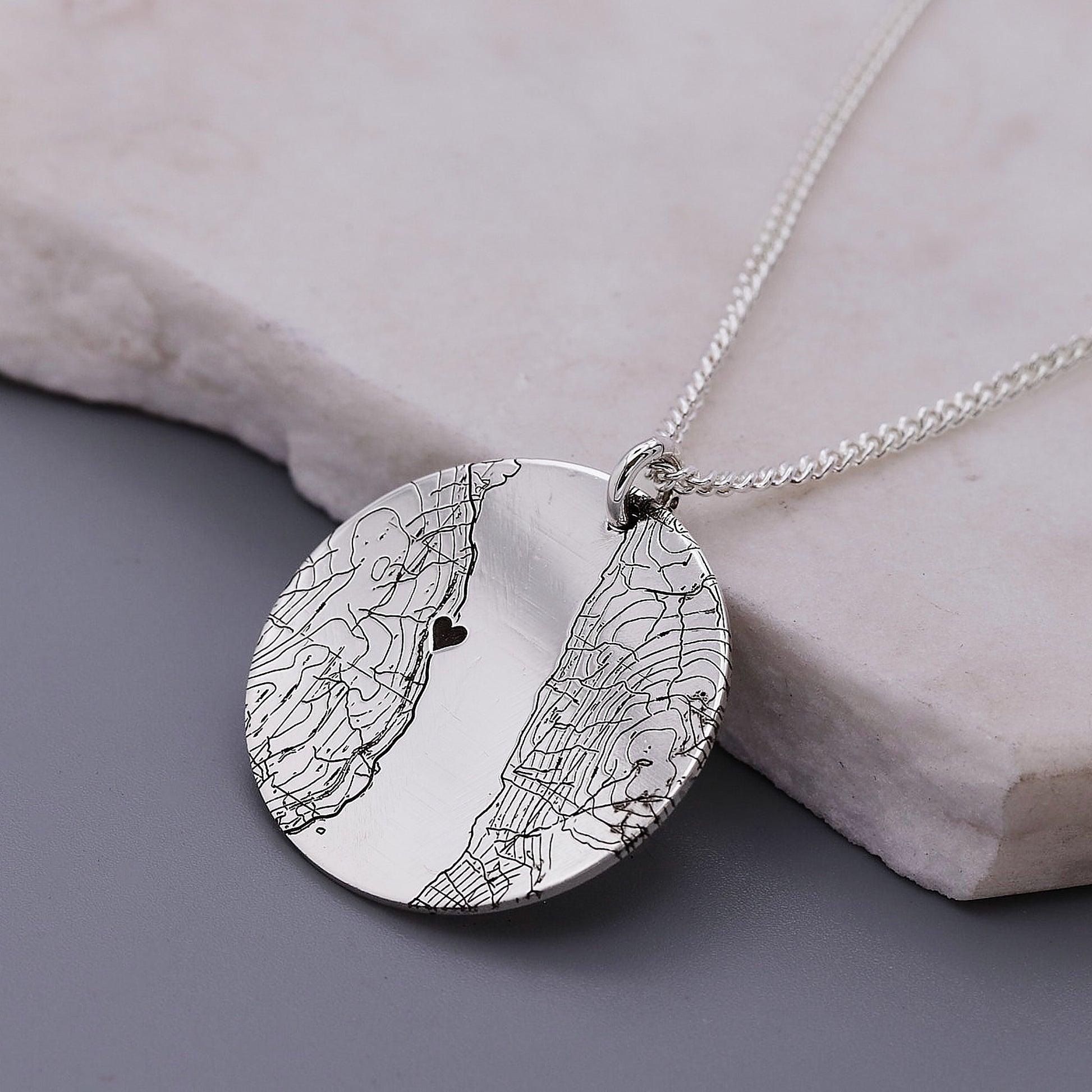 Silver necklace with a round pendant featuring a map design and a heart on a stone surface.