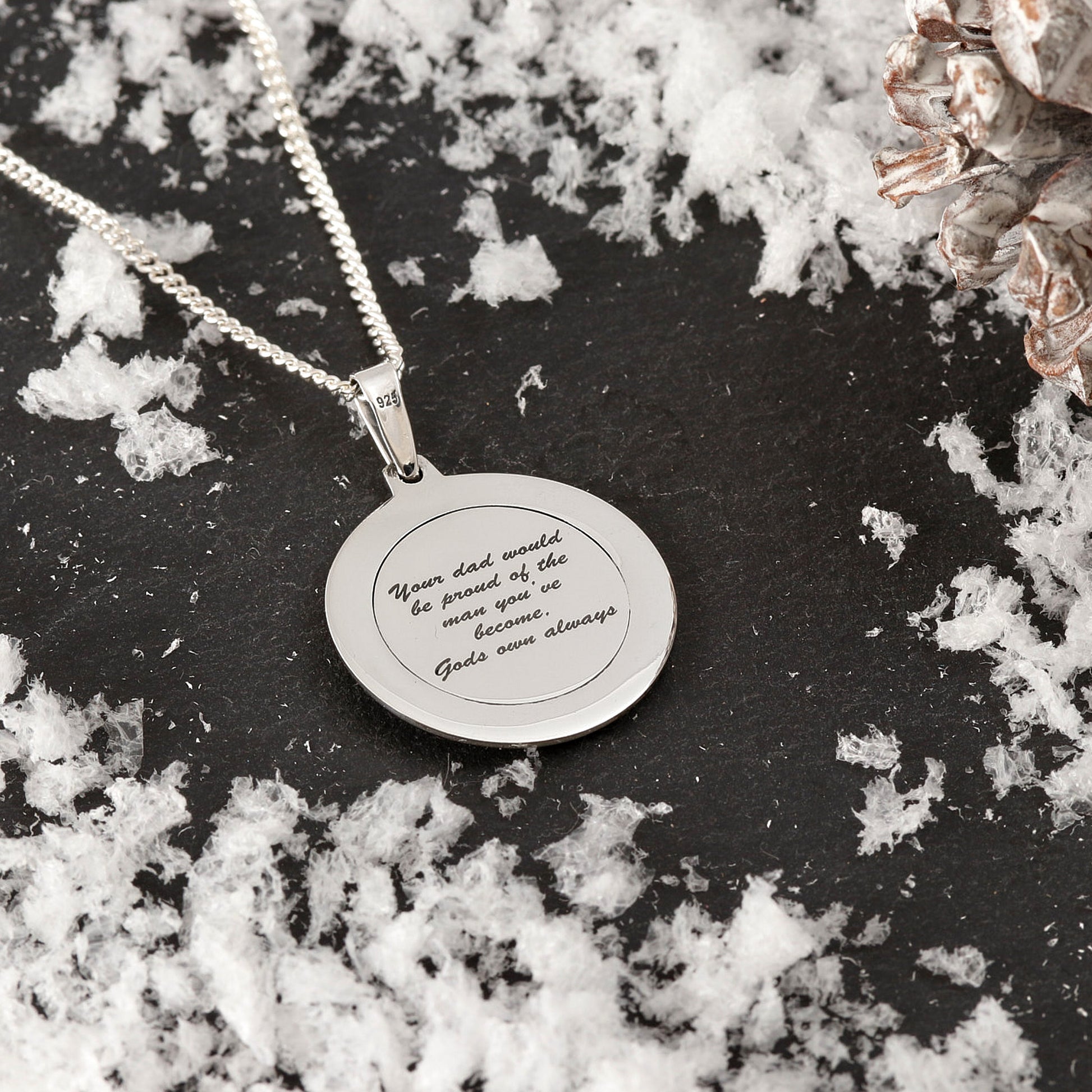 Silver necklace with a engraved pendant on a dark surface with white flowers.