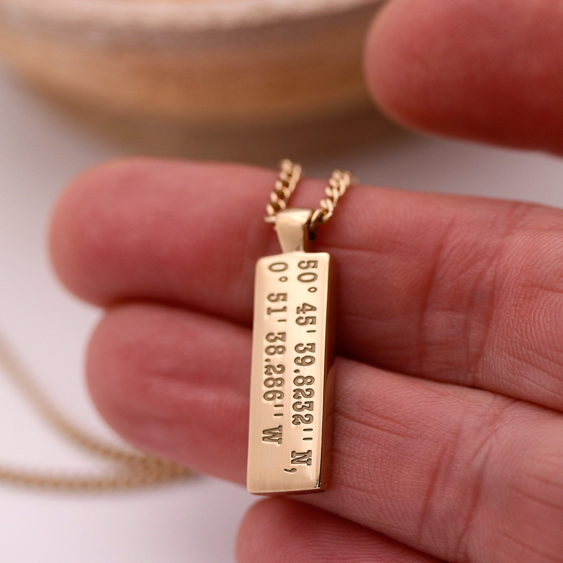 Gold necklace with a rectangular pendant held in a hand against a neutral background