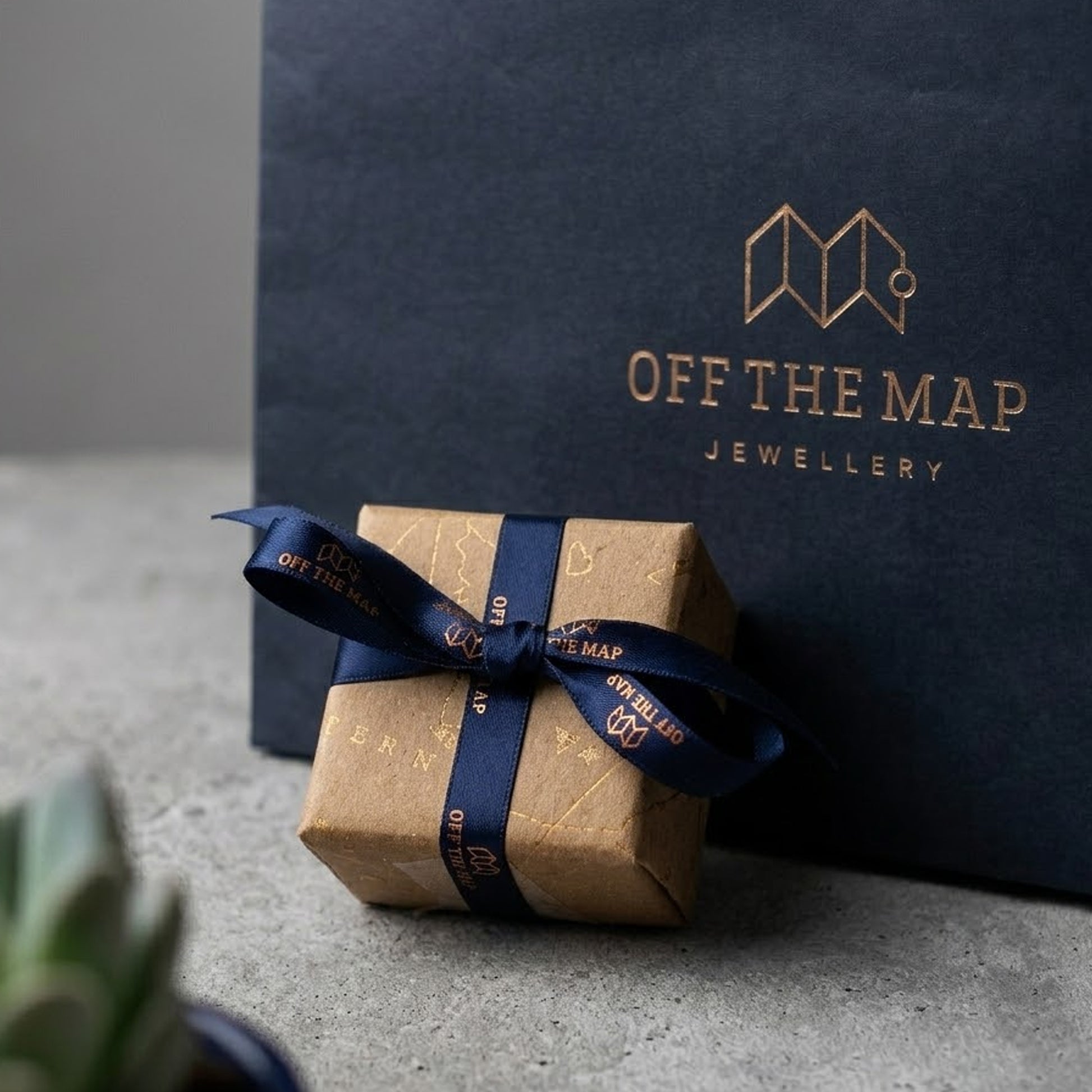 Gift box with blue ribbon and 'Off The Map Jewellery' branding in front of a branded box.