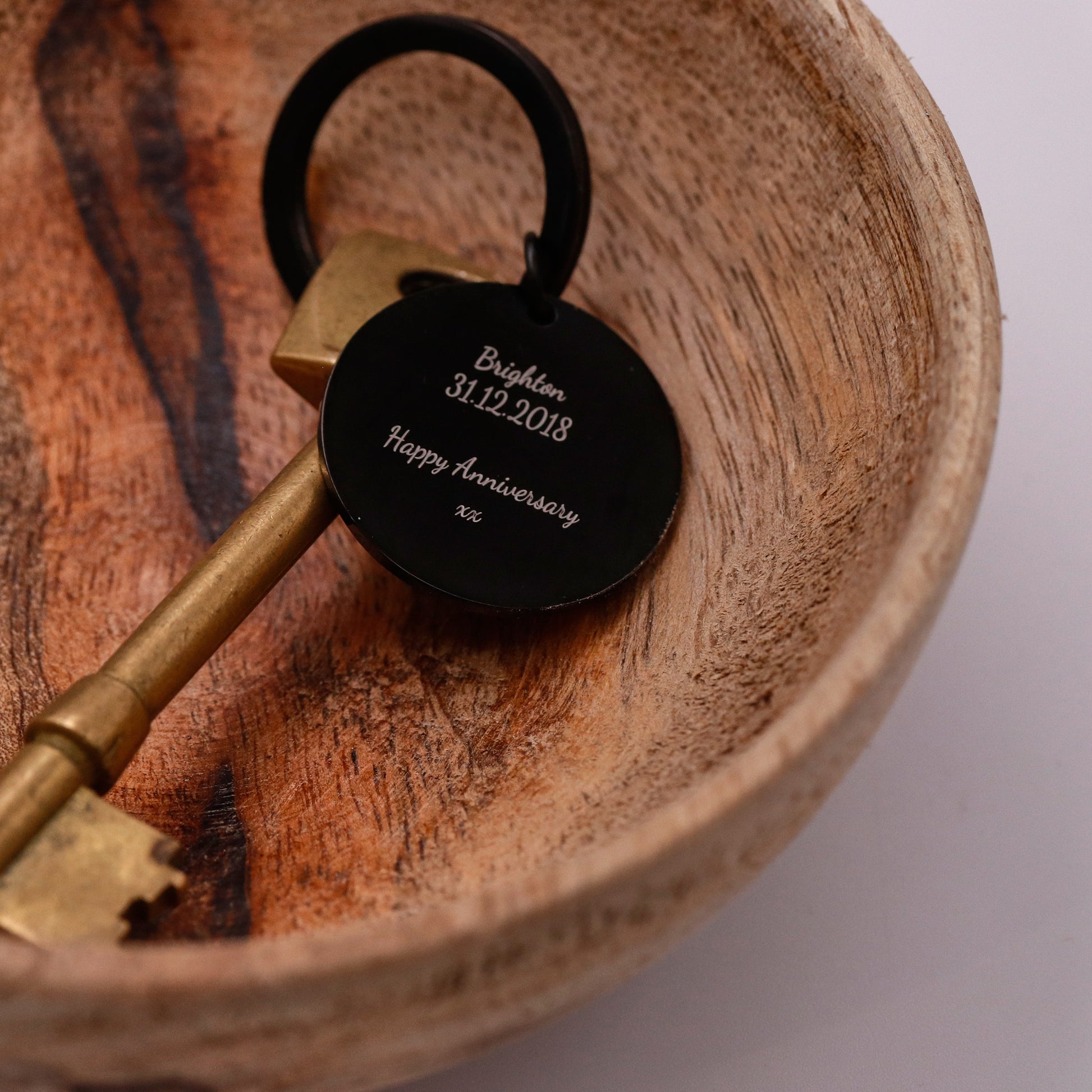 Bronze key with a black tag engraved with 'Brighton 31.12.2018 Happy Anniversary' on a wooden surface.