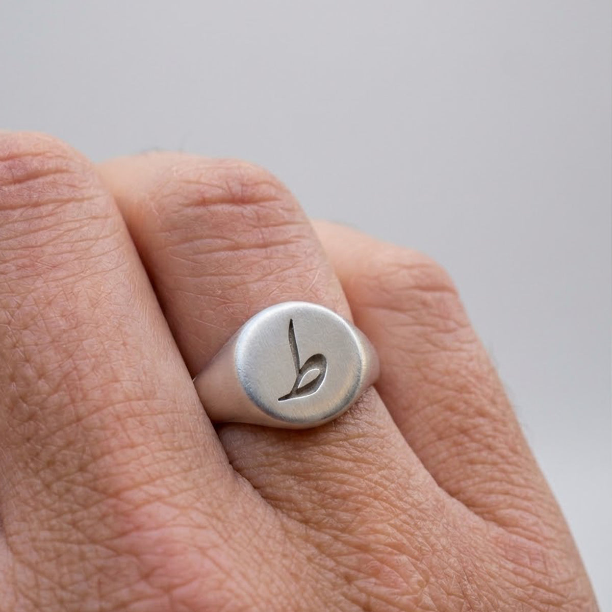 A silver signet ring with a stylized lowercase 'b' engraving, shown worn on a finger against a soft gray background.