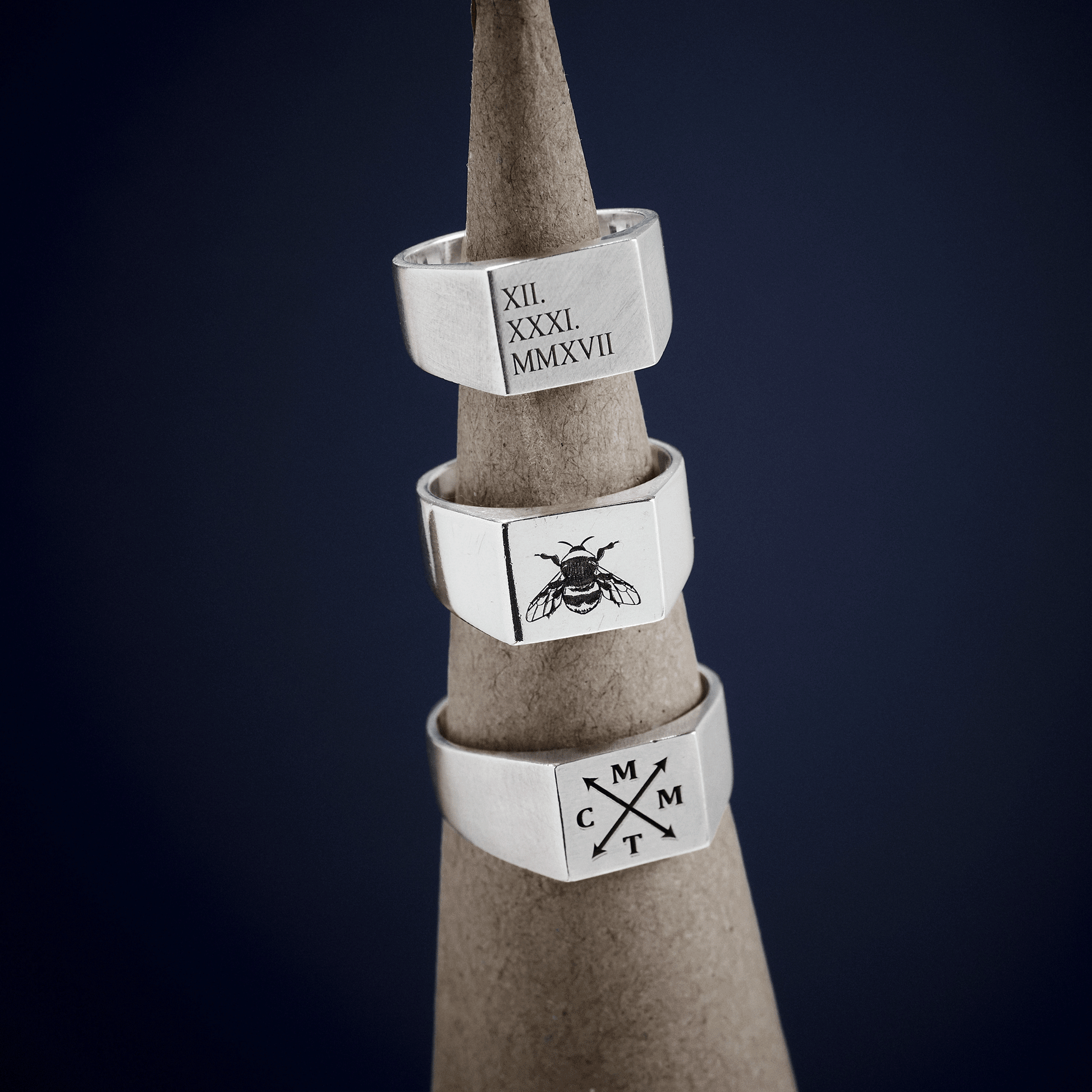 Three silver signet rings with custom engravings on a gray stand against a dark background