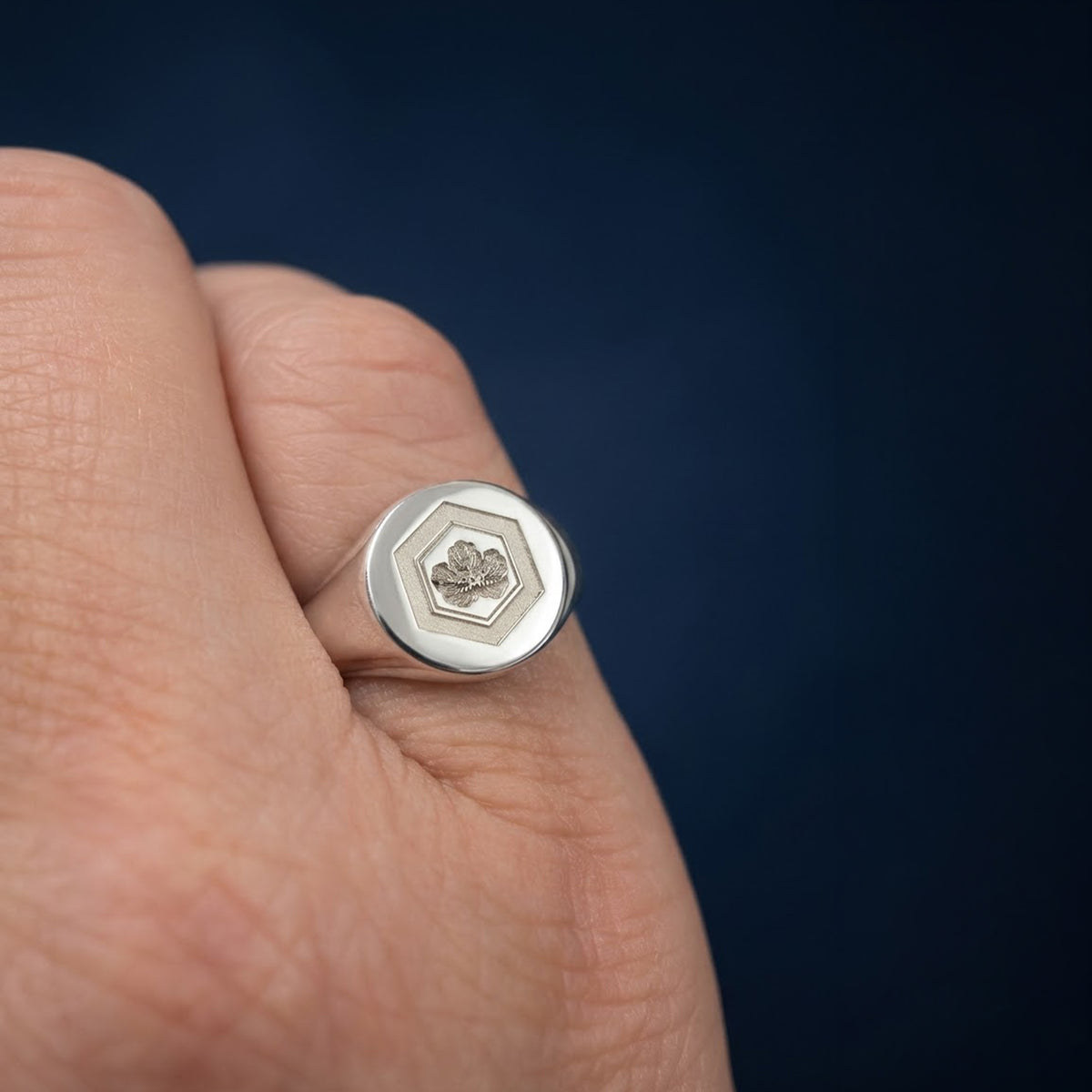 A silver signet ring with a hexagonal face and a small engraved floral motif, shown worn on a person's finger against a dark navy background.