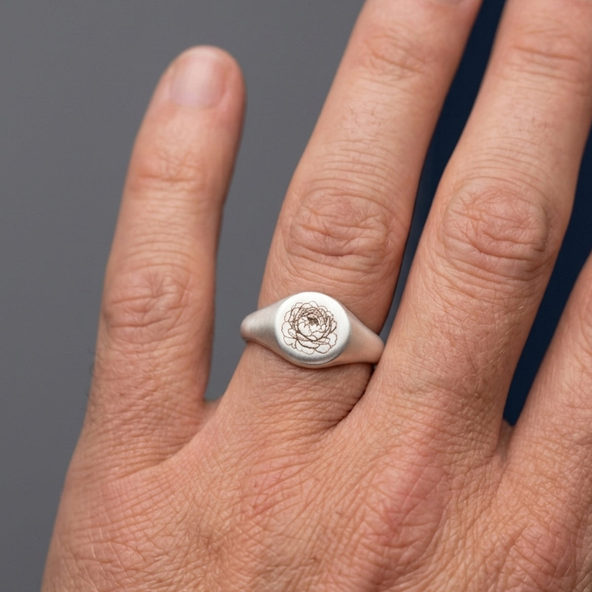 A silver signet ring with an engraved rose design on the face, shown worn on a person's ring finger against a dark gray background.