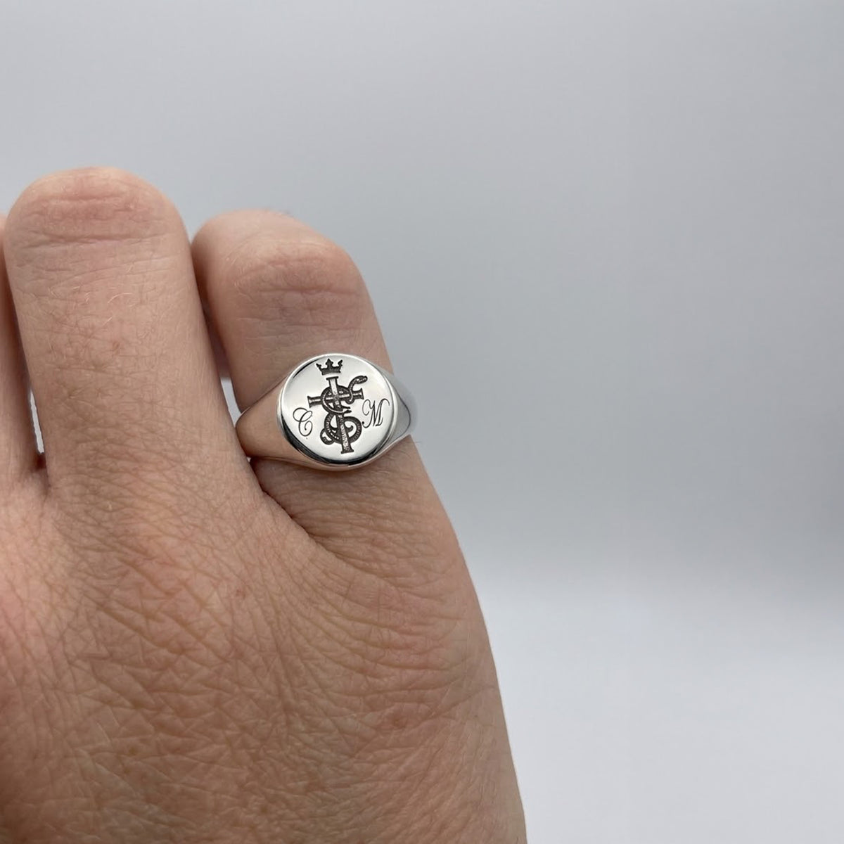 A silver signet ring with an engraved monogram and crown design, shown worn on a person's ring finger against a neutral gray background.