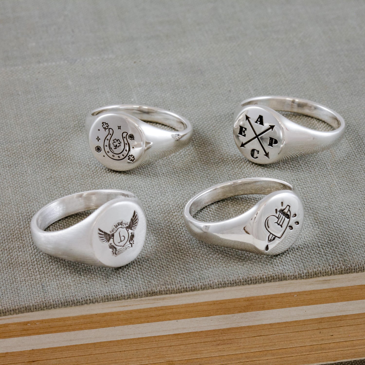 Four silver signet rings with engraved designs on a textured surface.