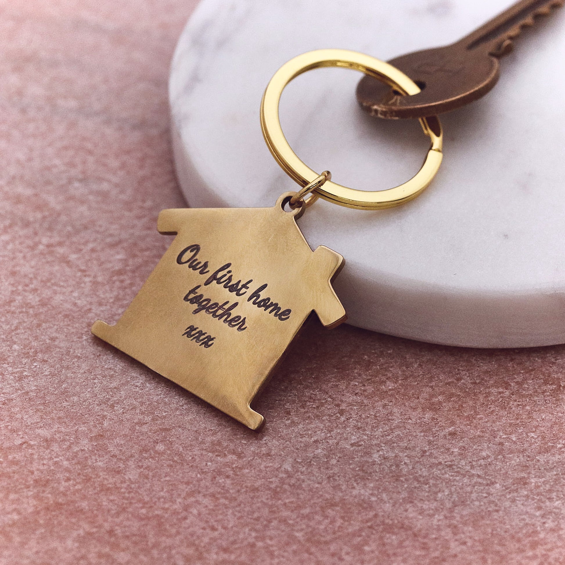 Gold keychain with 'Our first home together' engraving on a pink surface.
