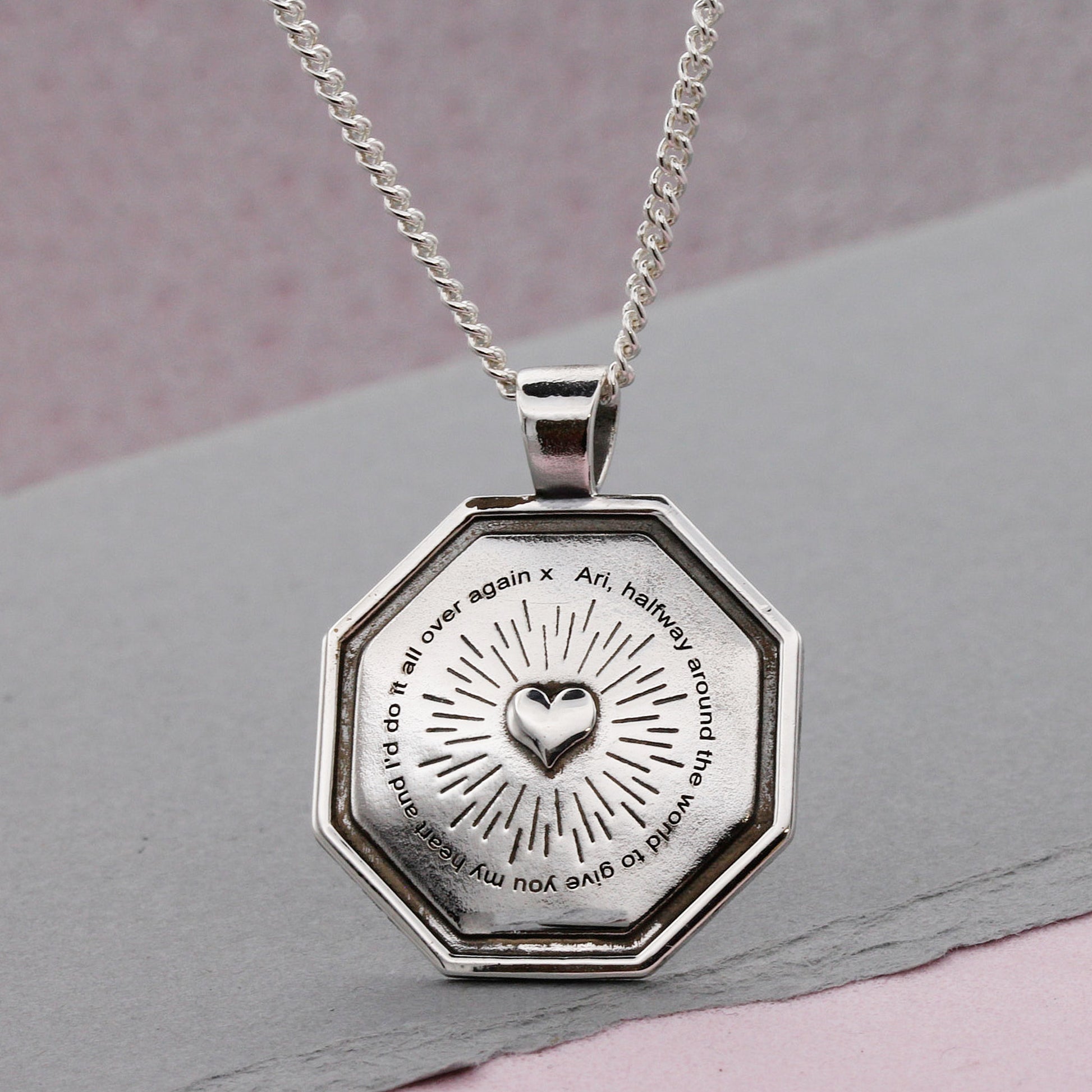 Silver octagonal pendant with engraved heart design and text on a chain against a gray background.