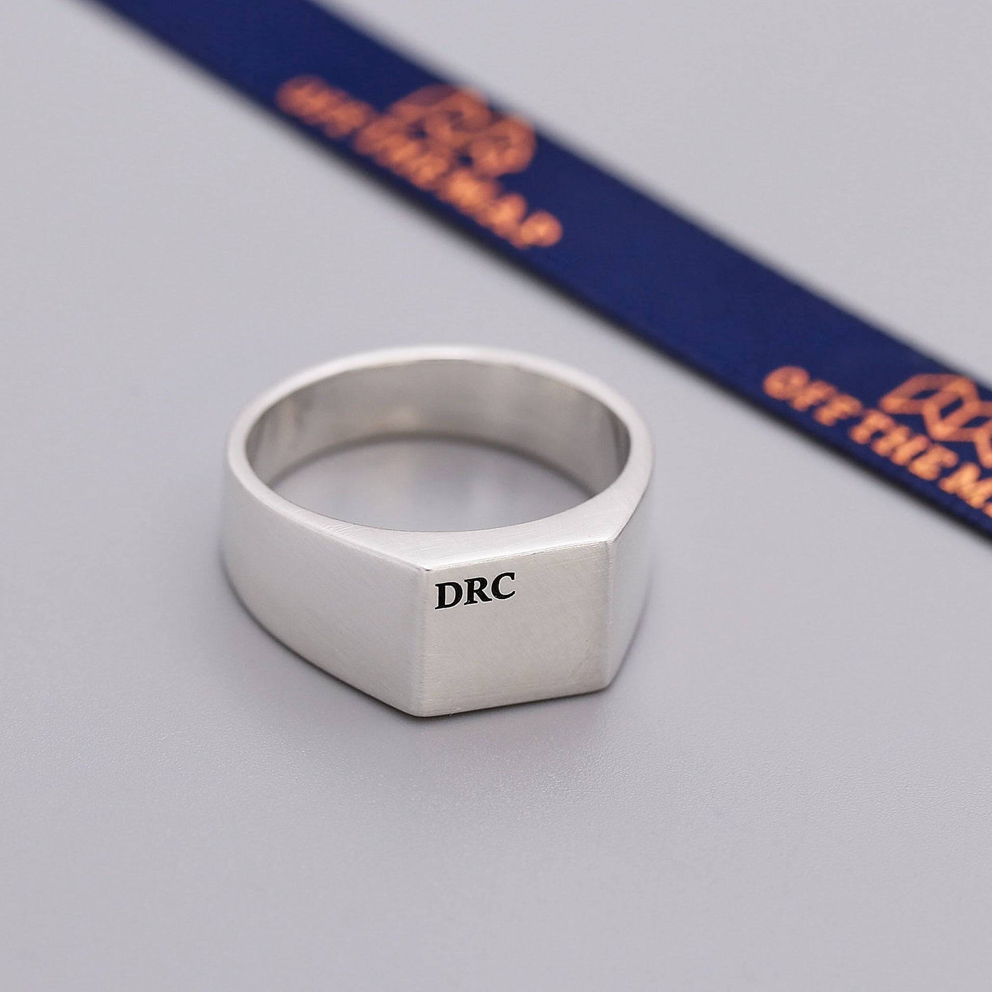 Silver ring with 'DRC' engraving on a gray surface with a blue and orange ribbon in the background.