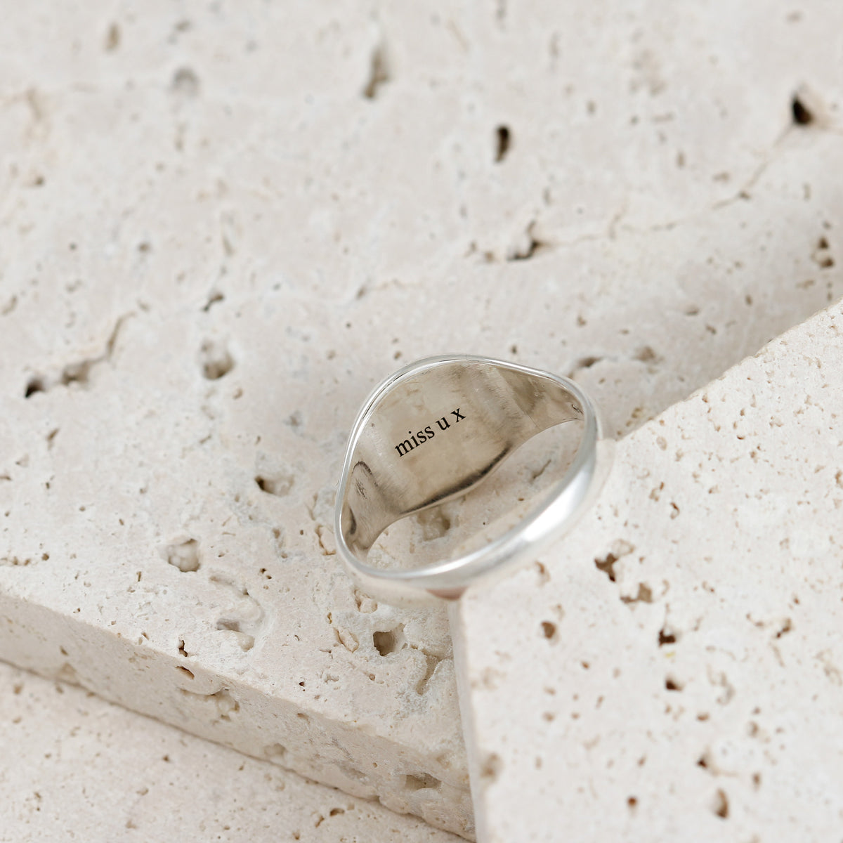 A polished silver ring with the engraved text 'miss u x' resting on a textured beige stone surface.