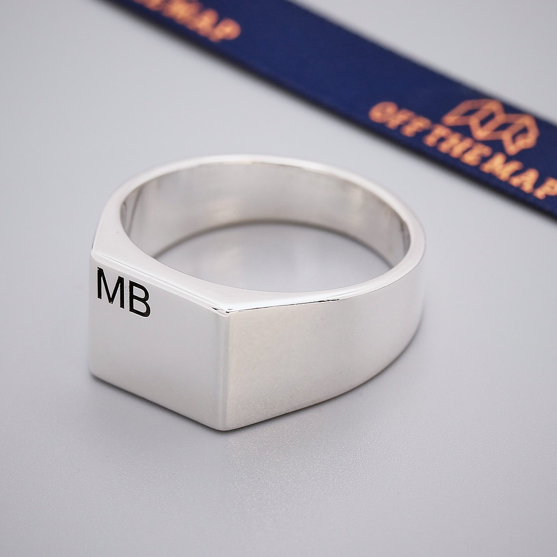 Silver ring with 'MB' engraving on a gray surface with a blue and orange strap in the background.