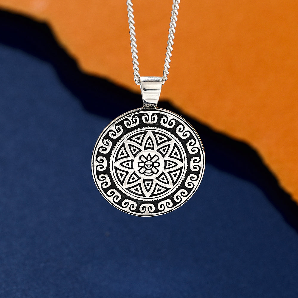 Silver necklace with a round, intricately designed aztec pendant on an orange and blue gradient background