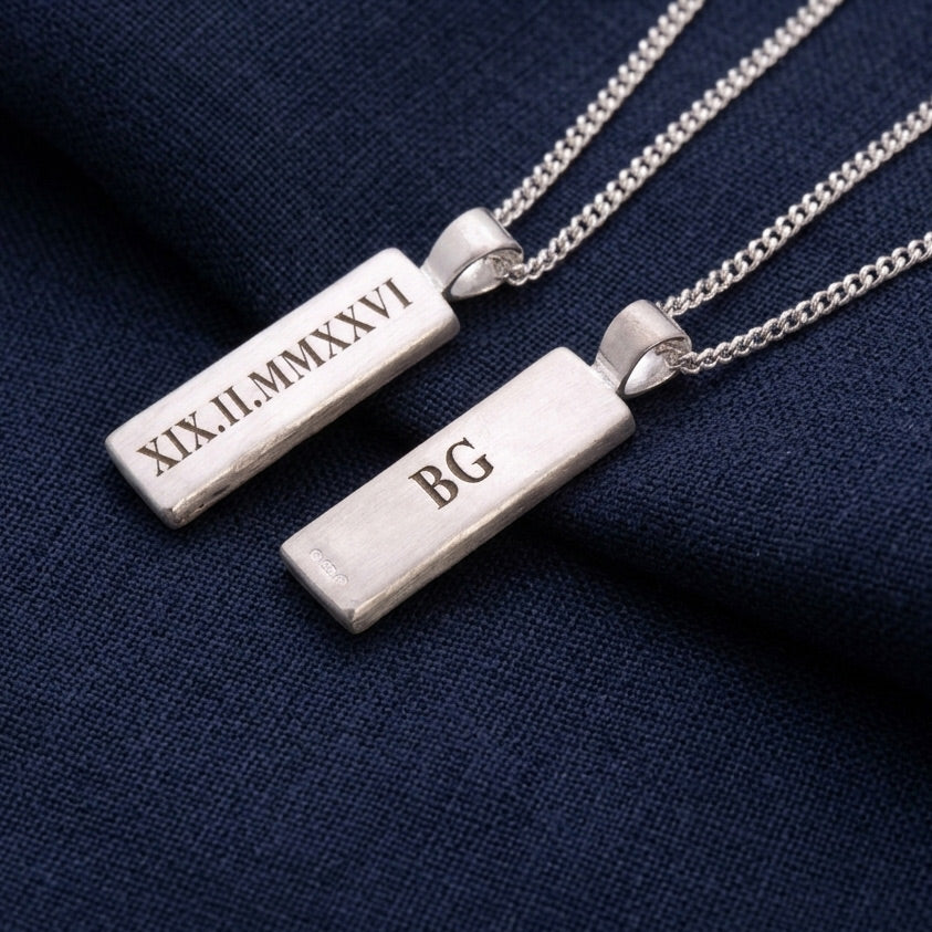 Two silver bar necklaces with engraved text on a dark blue fabric background