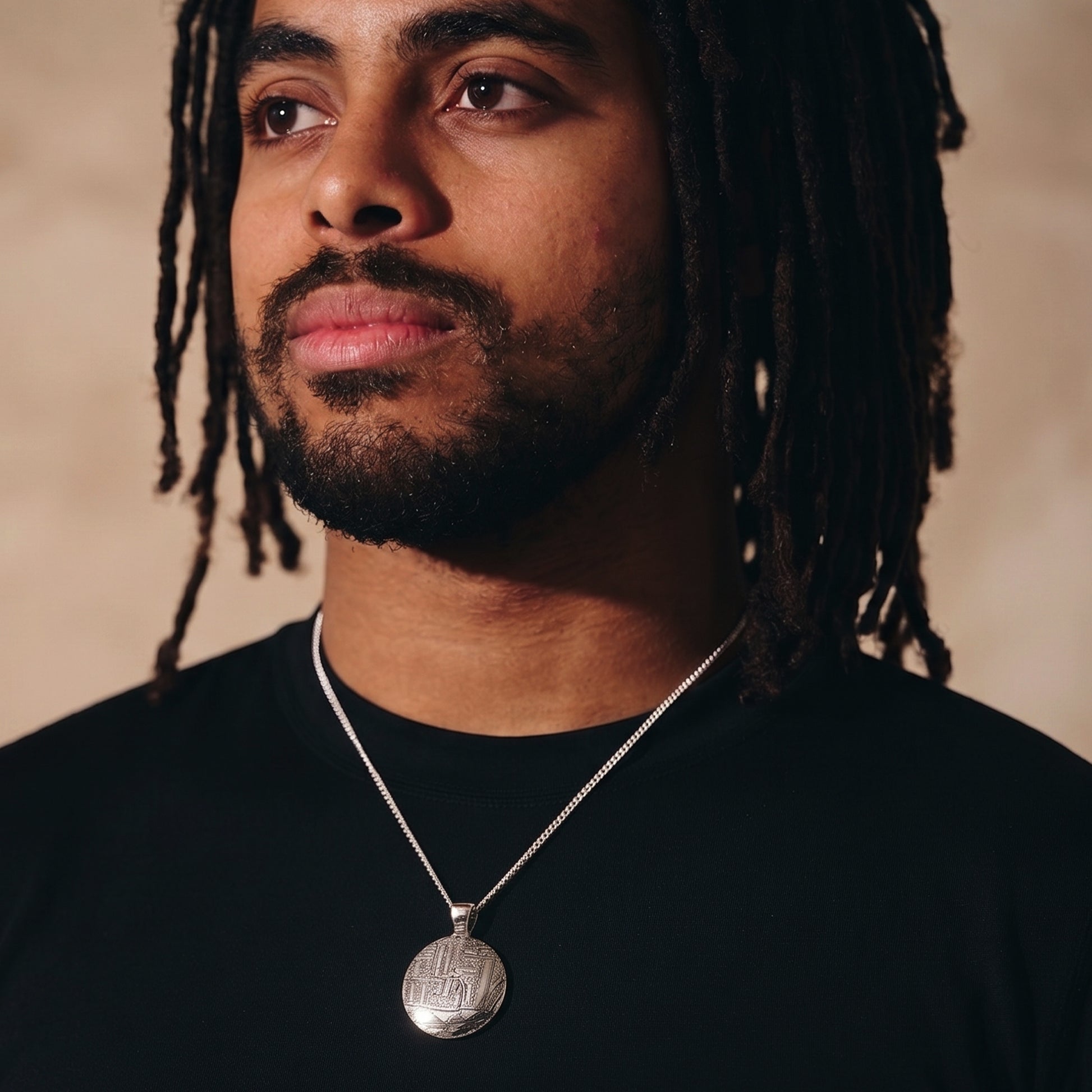Man with dreadlocks wearing a black shirt and necklace against a beige background