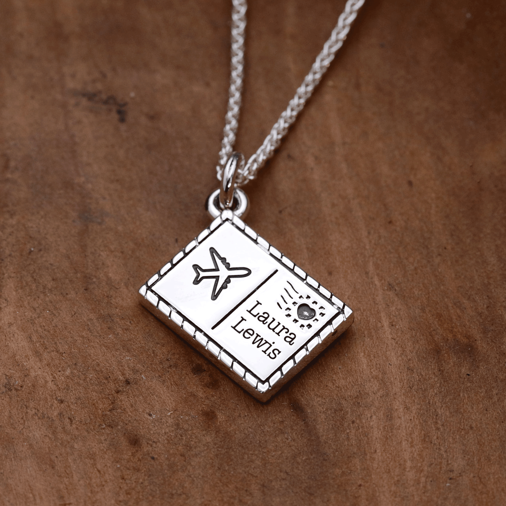 Silver necklace with a postcard pendant featuring engraved designs and text on a wooden surface