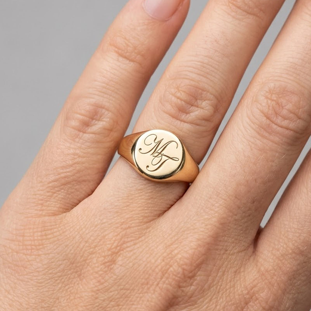 A close-up of a gold signet ring with an engraved monogram on a person's ring finger, shown against a neutral gray background.